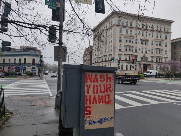Eine belebte Stadtstraße mit fahrenden Fahrzeugen, ein Fußgänger auf dem Gehweg, ein Müllcontainer mit der Aufschrift "Hände Waschen", Ampeln, Straßenschilder, ein Metallzaun, Grünfläche, Gebäude und einen klaren Himmel.