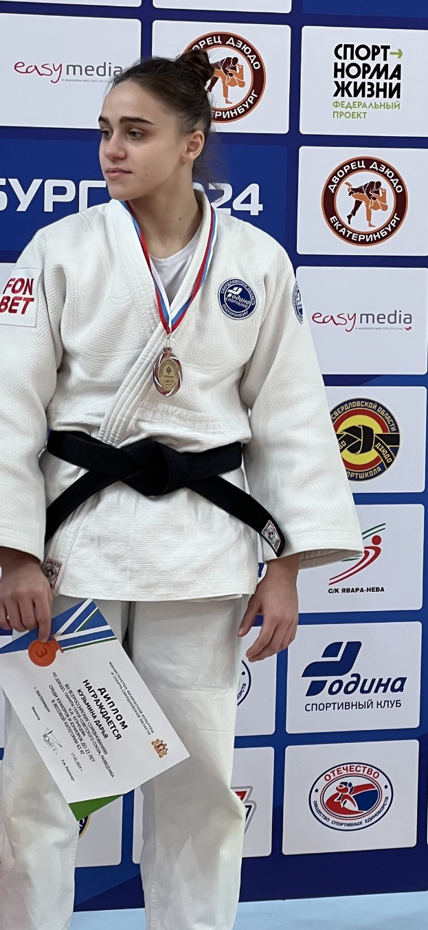 Eine Frau in einem weißen Judo-Anzug mit einem schwarzen Gürtel steht auf einem Podium und hält eine Medaille und ein Zertifikat in den Händen, mit einem logo-bedeckten Board im Hintergrund.