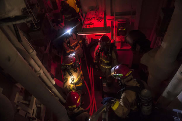 Feuerwehrleute in Helmen und Sauerstoffflaschen stehen in einem dunklen Raum mit Rohren an den Seiten, bereit für einen Rettungseinsatz in der Nähe einer Maschine und anderer Gegenstände.
