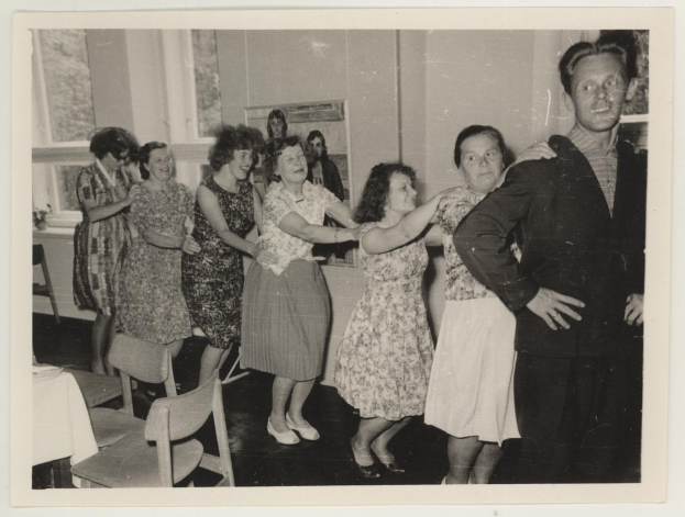 Gruppe von Menschen, die in einem Raum mit Stühlen und einem mit einer wei├čen Decke bedeckten Tisch tanzen. An der Wand befindet sich ein Foto und im Hintergrund sind Fenster zu sehen.