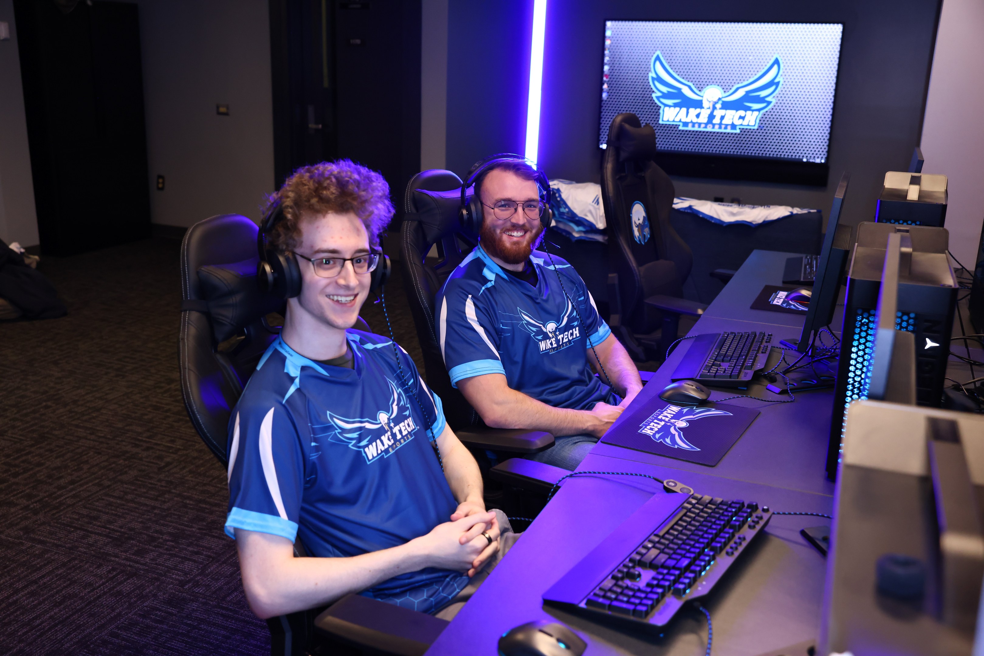 Zwei Männer mit Headsets sitzen an einem Schreibtisch mit Monitoren, Tastaturen und anderem Equipment und lächeln beim Spielen eines Spiels.