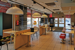 Das Innere eines Fast-Food-Restaurants mit Tischen und Stühlen, Hockern, einem Boden, einer Decke mit Lampen, einer Wand mit Feuerlöscher, Glastüren und diversen anderen Gegenständen.