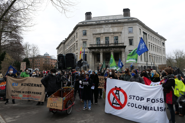 Große Gruppe von Menschen bei einer Demonstration gegen fossile Brennstoffe, die Schilder und Fahnen tragen, mit einem Fahrzeug im Vordergrund, Bäumen und Gebäuden im Hintergrund unter einem klaren blauen Himmel.