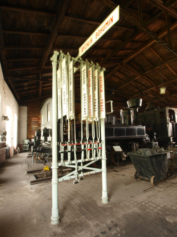 Ein Bahnsteig mit einem Zug auf den Schienen, einem Rollstuhl auf dem Boden, einer Signalbox in der Mitte, einem Pfahl mit einer Tafel, einer Wand links und einer Decke darüber.