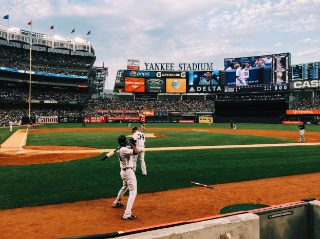 Baseball-Spiel im Yankee Stadium mit Spielern auf dem Feld und Zuschauern in den Rängen unter einem bewölkten Himmel.