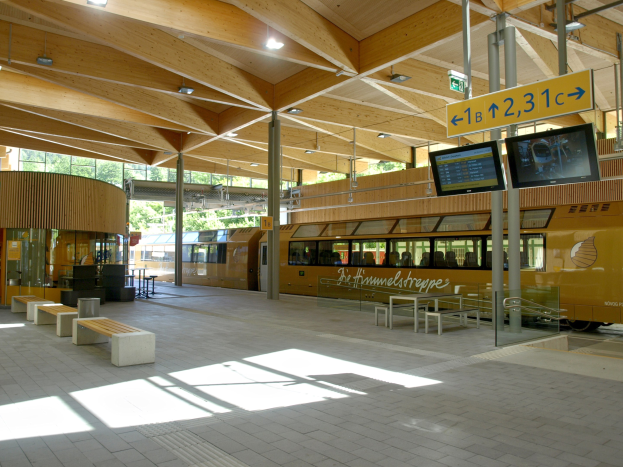 Ein belebter Bahnhof mit einem stehenden Zug auf den Gleisen, umgeben von Bänken, Tischen, Pfosten, Schildern, Laternen und Bäumen, während Menschen auf den Zug warten.
