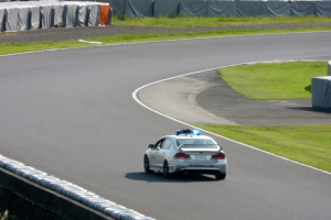 Ein Polizeiwagen fährt auf einer Rennstrecke mit Gras an beiden Seiten, mit Menschen, Zelten und anderen Gegenständen im Hintergrund.