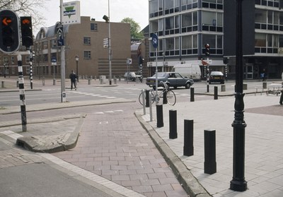Eine Stadtstraße mit einem geparkten Fahrrad, fahrenden Fahrzeugen, Fußgängern auf dem Gehweg, Versorgungsmasten, Verkehrsampeln, Schildern, Bäumen, Gebäuden mit Fenstern und einem klaren blauen Himmel.