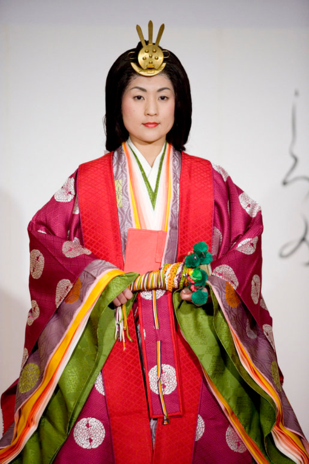 Eine Frau in einem traditionellen japanischen Kimono, das ein leuchtendes rotes und grünes Kleid mit aufwändigen Mustern zeigt, steht selbstbewusst auf einem Laufsteg, ihr Haar ist ordentlich zu einem Knoten gebunden.