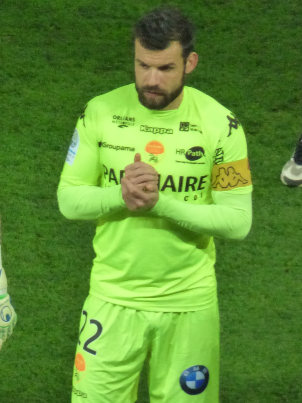 Ein Torwart in einem gelben Trikot klatscht auf einem Fußballfeld, trägt ein grünes Hemd und Shorts und hat einen Teil des Beins einer anderen Person auf der linken Seite sichtbar.