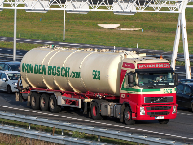 Großer weißer, roter und grüner Tanklastwagen fährt auf einer Autobahn mit anderen Fahrzeugen, einer Straßenbefestigung, Gras und einem Eisenrahmengebilde im Hintergrund.