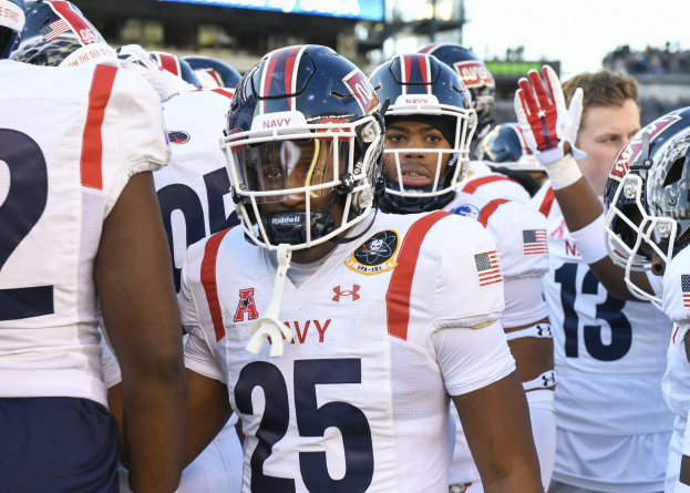 Gruppe von Männern in Fußballausrüstung auf einem Feld mit Helmen und Handschuhen, mit einer unscharfen Menge, einem Anzeigebrett mit 'Auburn vs Virginia Tech - 11/28/15 College Football Pick, Odds, and Prediction' und Himmel im Hintergrund.