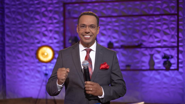 Stephen Colbert, Moderator von *The Daily Show with Trevor Noah*, steht auf einer Bühne und hält ein Mikrofon in der Hand, trägt einen Anzug, Krawatte und Brille, während im Hintergrund ein Scheinwerfer und ein Ständer zu sehen sind.