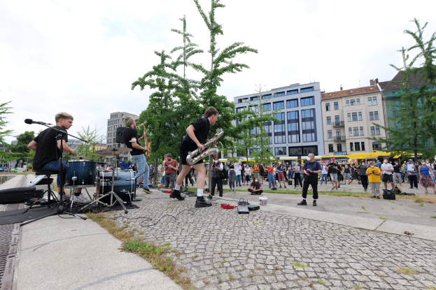 Eine Gruppe von Menschen, die Musik auf einem Bürgersteig mit Mikrofonen, Ständern und Equipment vor einem Publikum spielt.