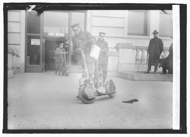 Schwarz-weißes Foto eines Mannes mit Mütze auf einem E-Scooter auf einem Gehweg, umgeben von anderen Menschen, darunter ein Radfahrer, und einem Gebäude mit einer Tür und einer Tafel im Hintergrund.
