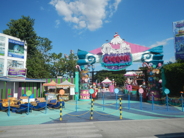 Vergnügungsfahrgeschäft am Parkeingang mit Straße, Umzäunung, Buden, Banner, Bäumen und bewölktem Himmel im Hintergrund.