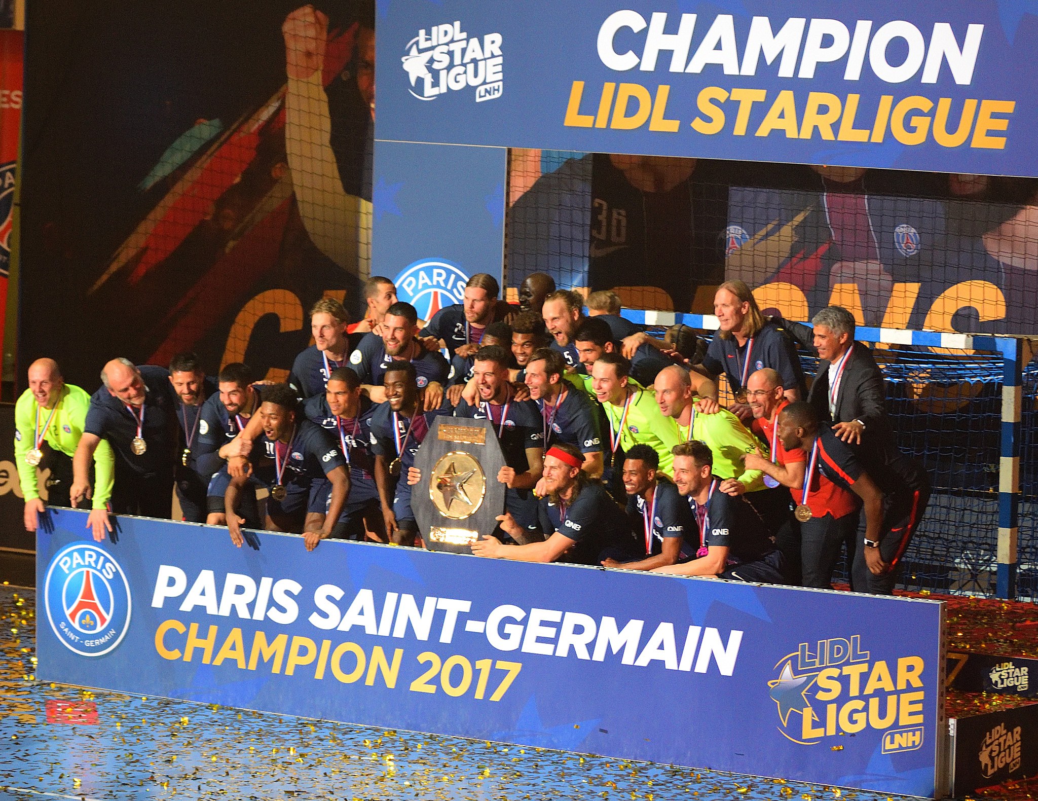 Spieler von Paris Saint-Germain feiern mit der Ligue-1-Trophäe, tragen Medaillen, vor einer Tafel mit Text und Logos, ein Netz rechts daneben und ein Bildschirm im Hintergrund.