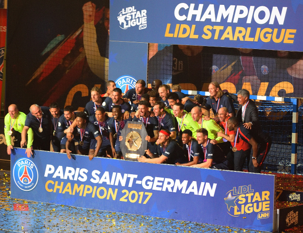 Spieler von Paris Saint-Germain feiern mit der Ligue-1-Trophäe, tragen Medaillen, vor einer Tafel mit Text und Logos, ein Netz rechts daneben und ein Bildschirm im Hintergrund.