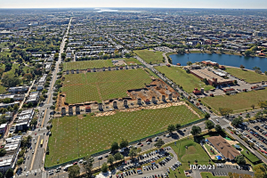 Luftaufnahme einer Stadt mit Gebäuden, Bäumen, Gras, Fahrzeugen, Wasser und Wolken, die einen großen zentralen Platz zeigt, der als Parkplatz vorgeschlagen ist.