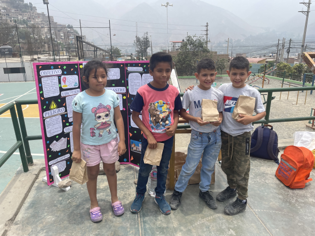 Gruppe von Kindern auf einem Basketballplatz mit Papiertüten in den Händen, daneben Tüten und Gegenstände auf dem Boden und eine Tafel mit Text links daneben, vor einer Kulisse aus Zäunen, Pfosten, Straßenlaternen, Strommasten, Bäumen, Gebäuden, Hügeln und Himmel.