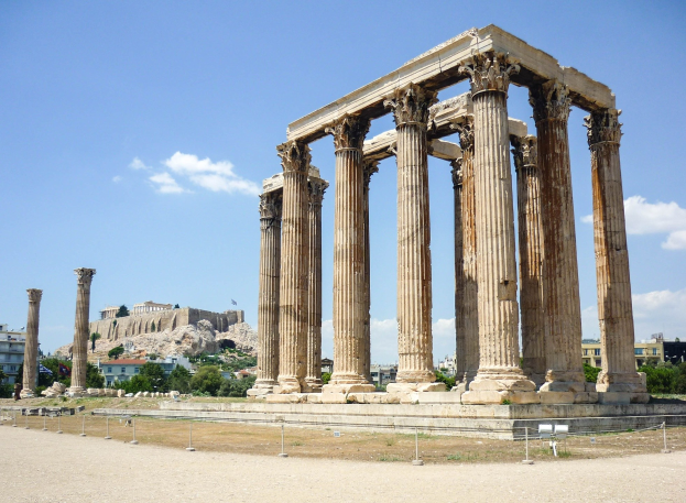 Alter Tempel von Olympian Zeus in Athen, Griechenland, mit seinen hohen korinthischen Säulen, umgeben von Bäumen, Felsen und einer Burg im Hintergrund unter einem bewölkten Himmel.