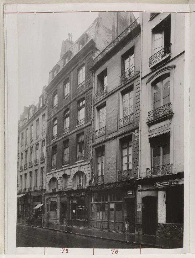 Schwarz-weißes Foto einer Stadtstra├če mit Geb├Ąuden, die Fenster, Gel├Ąnder und Namensschilder aufweisen, und einer Stra├če unten.