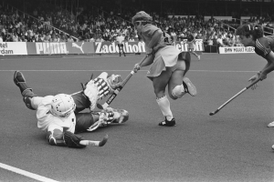 Schwarzes und weißes Foto von Frauen, die Feldhockey in einem Stadion mit Zuschauern in den Rängen spielen, Helme tragen und Stöcke halten, umgeben von Tafeln mit Text.