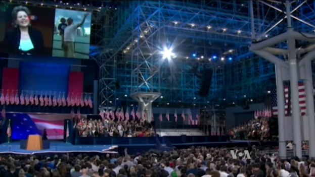 Eine große Menschenmenge sitzt vor einer Bühne mit einem Podium, einem Mikrofon, Flaggen, einem Bildschirm und Lichtausrüstung und wartet auf eine Präsidentenrede.
