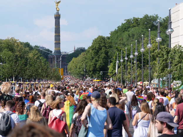Eine große Menschenmenge geht eine Straße in Berlin, Deutschland, entlang, mit Laternen, Bäumen und einem Turm mit einer Statue im Hintergrund und Gebäuden unter einem bewölkten Himmel.