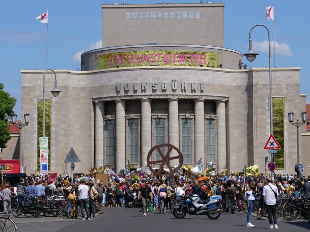 Große Gruppe von Menschen versammelt sich friedlich vor einem Gebäude mit Säulen und Text in Berlin, Deutschland, mit Straßeninfrastruktur, Fahrzeugen, Bäumen und bewölktem Himmel im Hintergrund.
