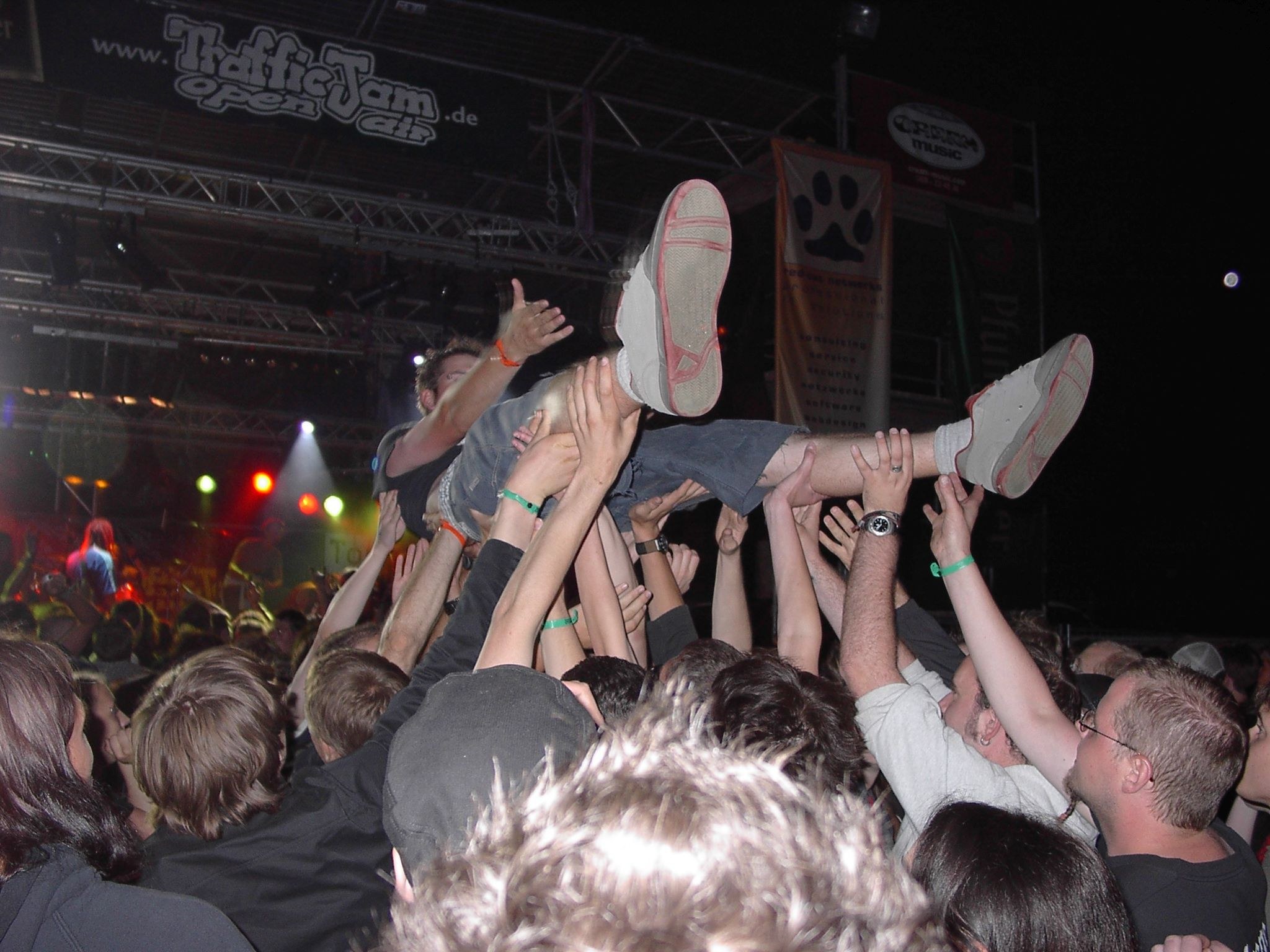 Menschenmenge auf einem Konzert mit erhobenen Händen, einige tragen Kappen, eine Person hält eine andere, unter Bühnenlicht und Trägern mit Bannern in einer dunklen Atmosphäre.