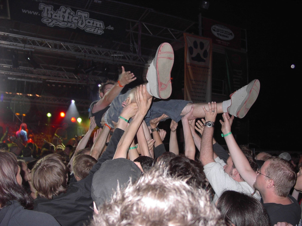 Menschenmenge auf einem Konzert mit erhobenen Händen, einige tragen Kappen, eine Person hält eine andere, unter Bühnenlicht und Trägern mit Bannern in einer dunklen Atmosphäre.