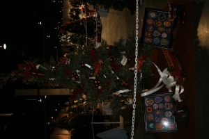 Ein Weihnachtsbaum mit Geschenken, Rahmen und Spielzeugen auf einer Plattform, mit Menschen, weiteren Weihnachtsb├Ąumen und Deko im Hintergrund.