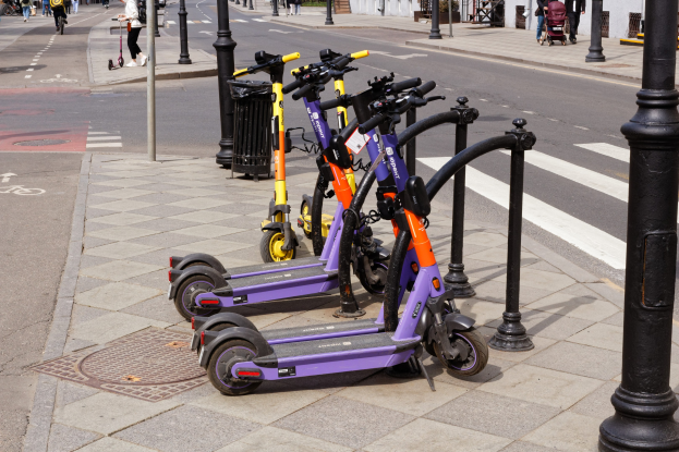 Eine Reihe von Elektro-Scootern, die an einer Straße geparkt sind, mit Fußgängern, Radfahrern, einem Müllcontainer im Vordergrund und Gebäuden, Bäumen und Pflanzen im Hintergrund.