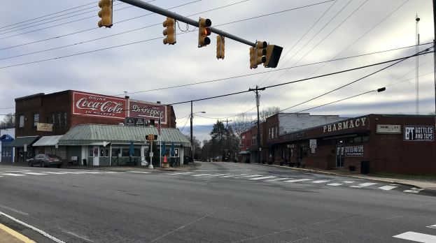 Stadtstraßenkreuzung mit einer Ampel, Fahrzeuge, Gebäude mit Namensschildern, Strommäste mit Drähten, Bäume und ein bewölkter Himmel.