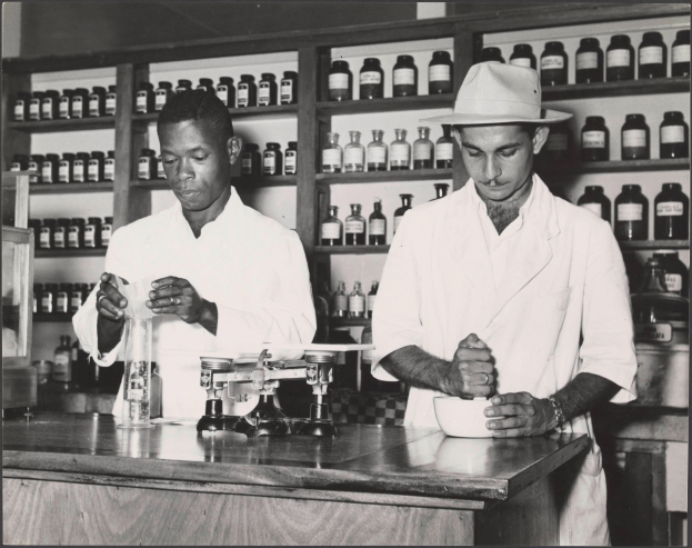 Zwei Männer in Laborkitteln und Hüten stehen an einem Tresen in einer Apotheke mit einer Waage, einer Schüssel und Regalen voller Flaschen im Hintergrund, dargestellt in Schwarz-Weiß.