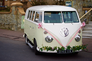 Ein weißer und rosafarbener VW-Bus, der mit Blumen und Blättern geschmückt ist, vor einer Steinwand geparkt, mit einer Treppe auf der rechten Seite und einem Gebäude im Hintergrund.