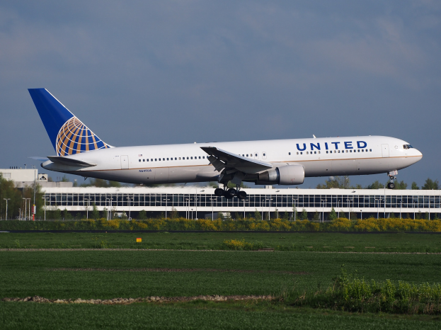 United Airlines Boeing 787-9 Dreamliner beim Abheben von einer Flughafenpiste mit saftig grünem Gras und Pflanzen, einem Gebäude und Bäumen im Hintergrund und einem klaren blauen Himmel darüber.