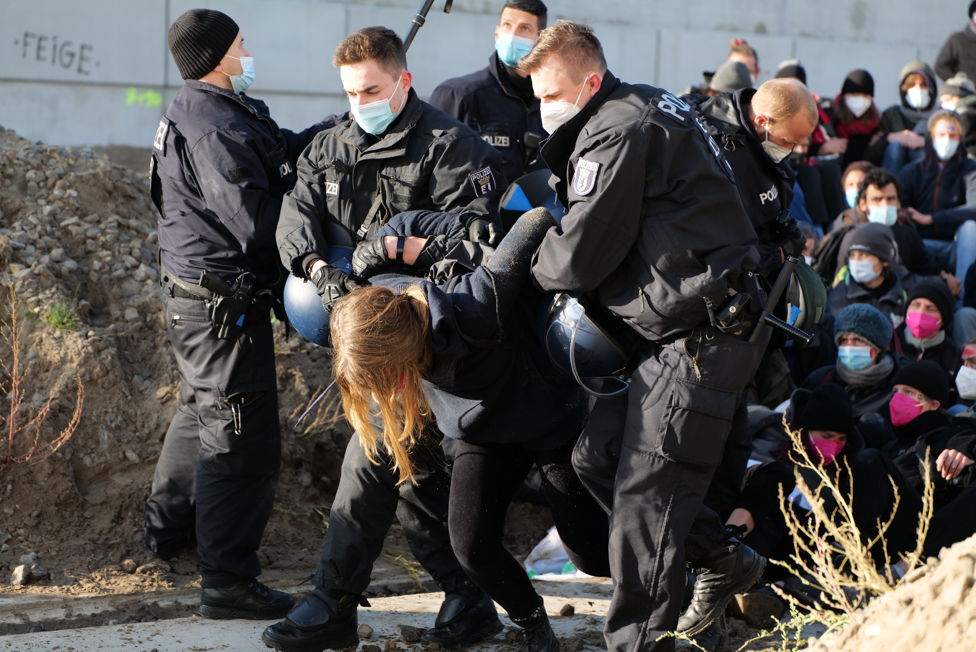 Eine Gruppe maskierter Personen in Mützen, einige bewaffnet, umgibt eine Frau, die von der Polizei weggeführt wird, mit Pflanzen, Steinen und einem protestbezogenen Wandtext im Hintergrund.