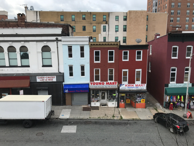Luftaufnahme einer Stadtstraße in Brooklyn, New York, mit Fahrzeugen, Geschäftsfronten, Werbetafeln, Strommasten und Drähten vor einem bewölkten Himmel, wobei ein kommerzielles Objekt zum Verkauf zu sehen ist.