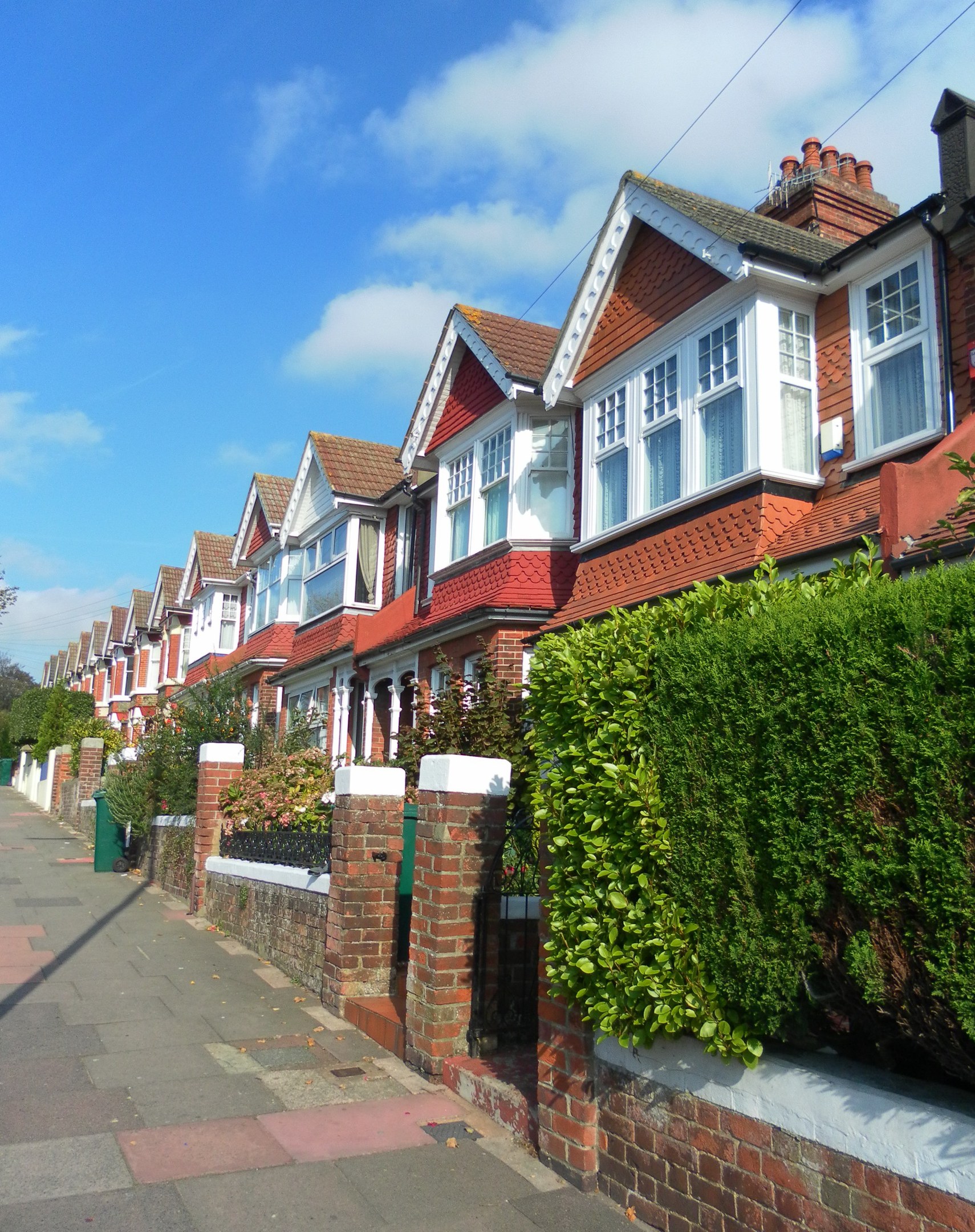 Reihe von Reihenh├Ąusern mit Glasfenstern, umgeben von B├Ąumen und Pflanzen, unter einem Himmel mit wei├čen Wolken in London.