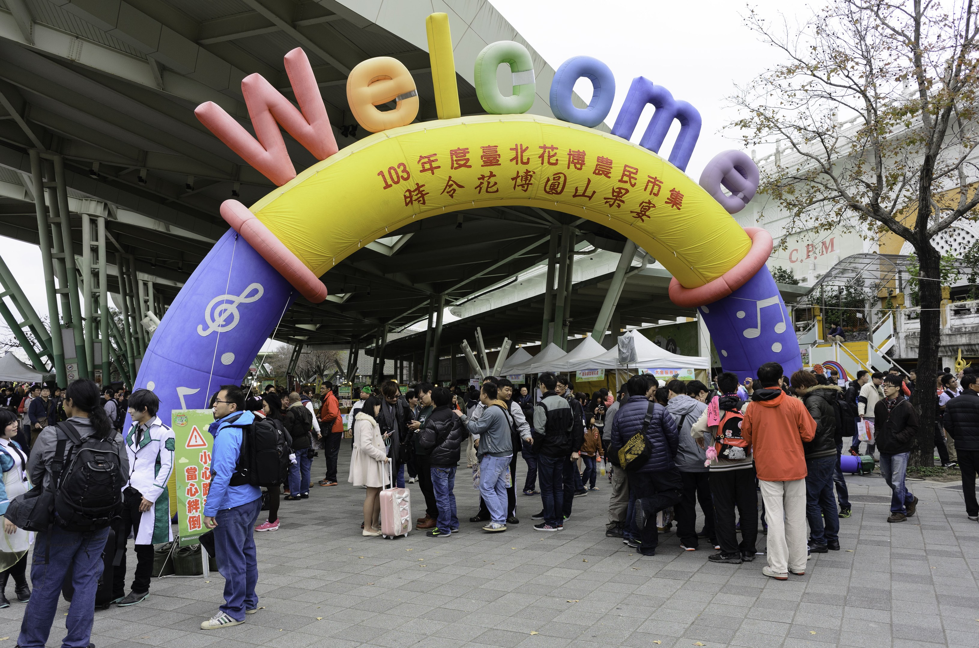 Eine Gruppe von Menschen steht vor einem Willkommensbogen bei einem Outdoor-Event mit Zelten, Bäumen und Gebäuden unter einem klaren blauen Himmel, einige tragen Taschen.