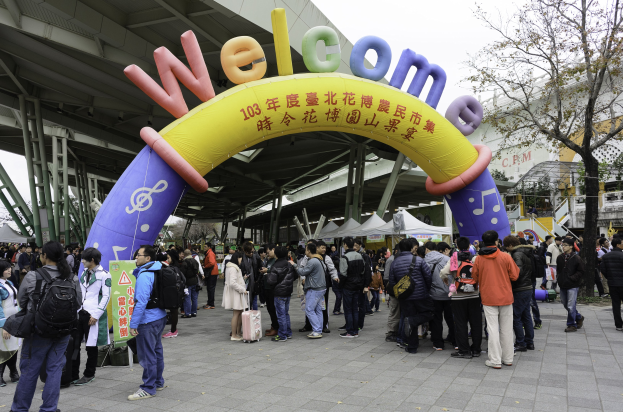 Eine Gruppe von Menschen steht vor einem Willkommensbogen bei einem Outdoor-Event mit Zelten, Bäumen und Gebäuden unter einem klaren blauen Himmel, einige tragen Taschen.
