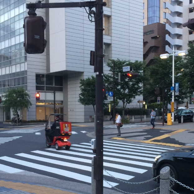 Ein E-Scooter auf einer Straße mit einer Ampel im Vordergrund, Personen im Hintergrund und Gebäude mit Bäumen dahinter.