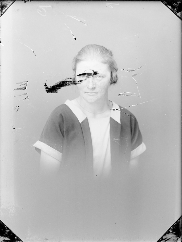 Ein Schwarz-Weiß-Foto einer Frau mit einem zerbrochenen Glasfenster im Hintergrund, die direkt in die Kamera schaut und ihre Hände vor der Brust verschränkt.