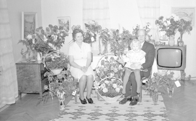 Schwarz-weißes Foto einer Familie aus drei Personen - ein Mann hält ein Kind, eine Frau und das Kind sitzen auf einem Sofa mit Blumenvasen auf den Seitentischen, einem Teppich auf dem Boden, Vorhängen im Hintergrund und Fotorahmen an der Wand.