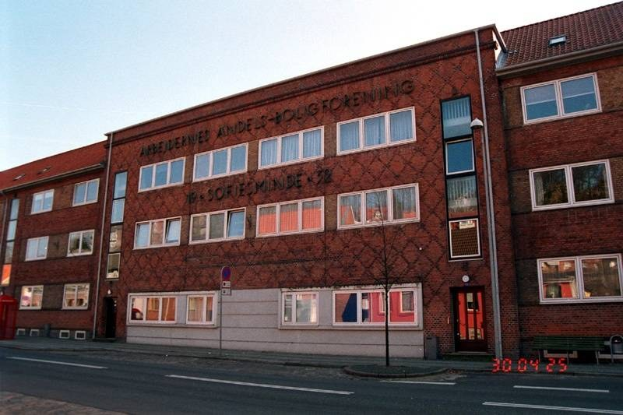 Großes Backsteingebäude mit vielen Fenstern, als deutsche Bundespolizei-Hauptquartier identifiziert, das sich auf einer Straße mit einem Gehweg, einem Baum, einer Bank, einem Laternenpfahl und einem roten Briefkasten befindet, vor einem Himmel als Hintergrund.