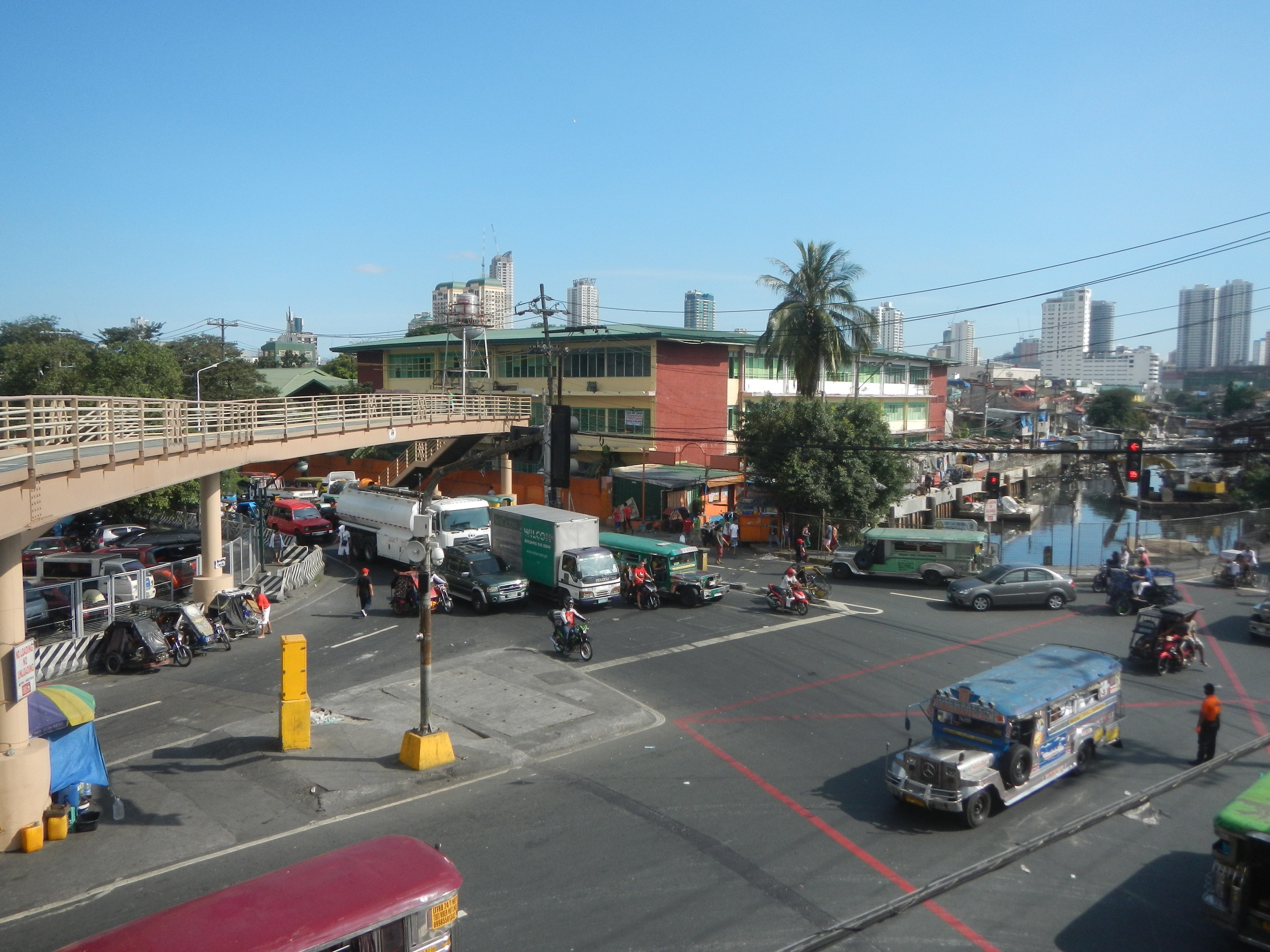 Lebendige Stadtstraße mit Autos, Bussen, Motorrädern, einer Brücke mit Geländern und Säulen, Laternen, Strommasten mit Drähten, Gebäuden mit Fenstern, Bäumen und einem bewölkten Himmel.