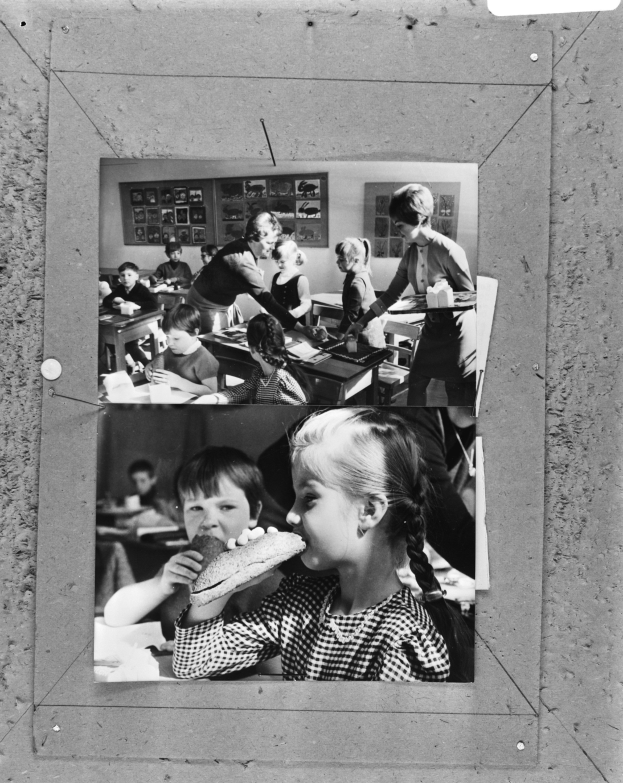 Ein junges Mädchen isst ein Sandwich in einem Klassenzimmer, mit anderen Kindern und Tischen mit Gegenständen im Hintergrund und Rahmen an der Wand.