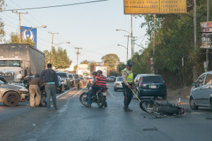 Eine Gruppe von Menschen steht um ein verunglücktes Motorrad auf der Seite einer Straße mit mehreren Fahrzeugen, darunter ein Lastwagen, und einer Hintergrund von Bäumen, Polen, Lampen und Schildern unter dem Himmel.